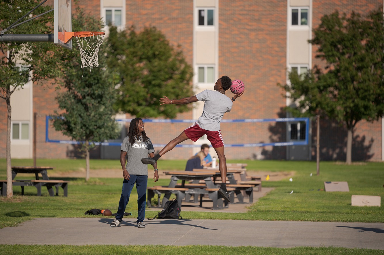 The image is of two students playing basketball. One student is jumping to dunk the basketball.