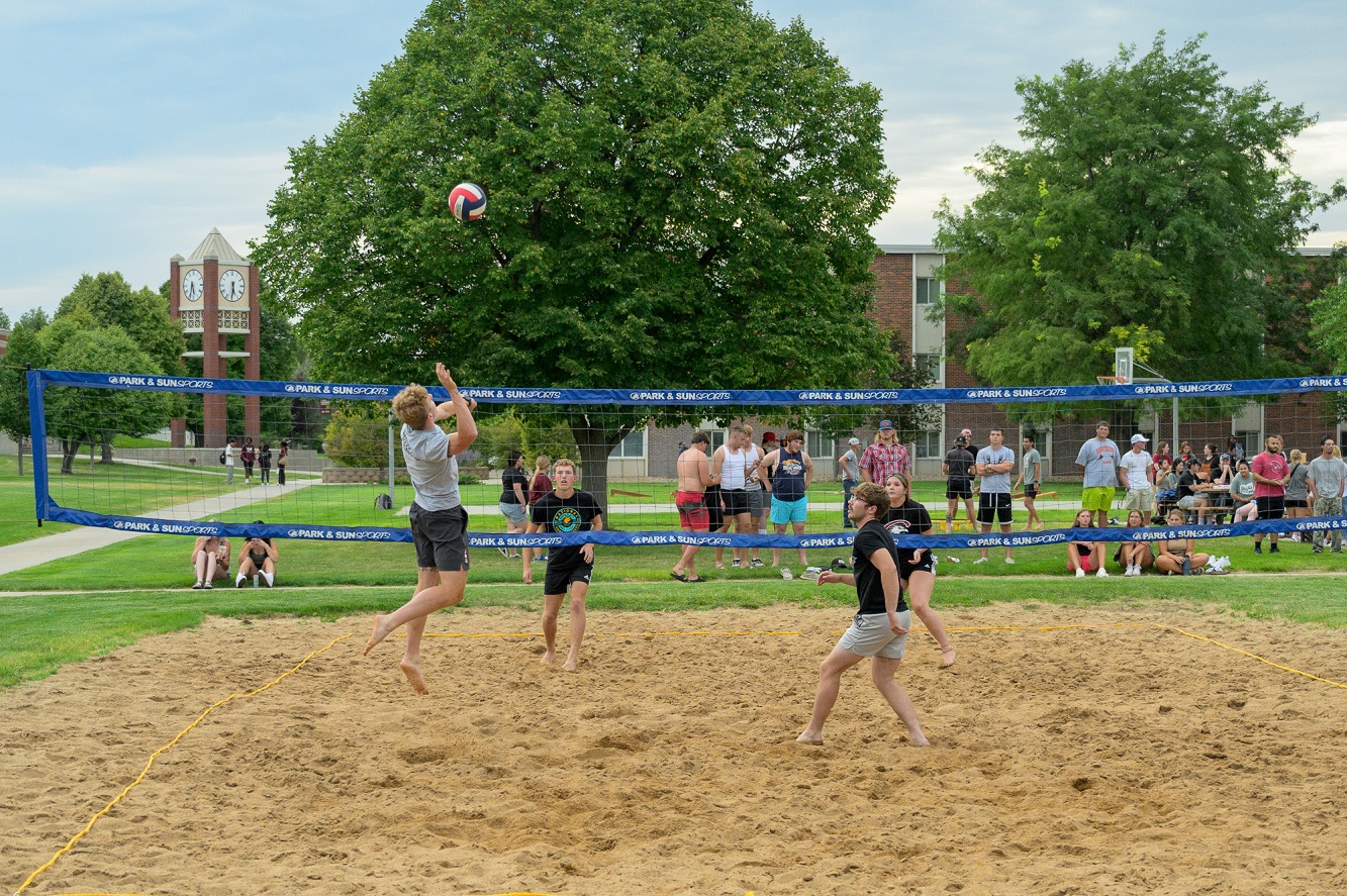 The image is of students playing volleyball with lots of green trees behind them.
