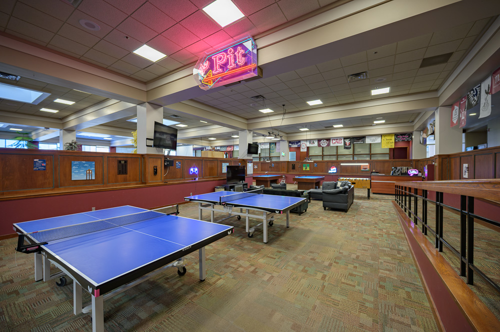 The image is of the Student Center area known as the Pit. To the right is an incline that leads to the front desk. There are two blue ping pong tables with several black couches and chairs. There is a foosball table next to two billiards tables in the back next to the front desk. There is also a large neon sign that reads 