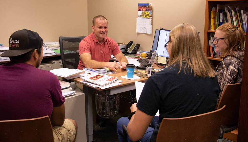Male professor at desk talking to students